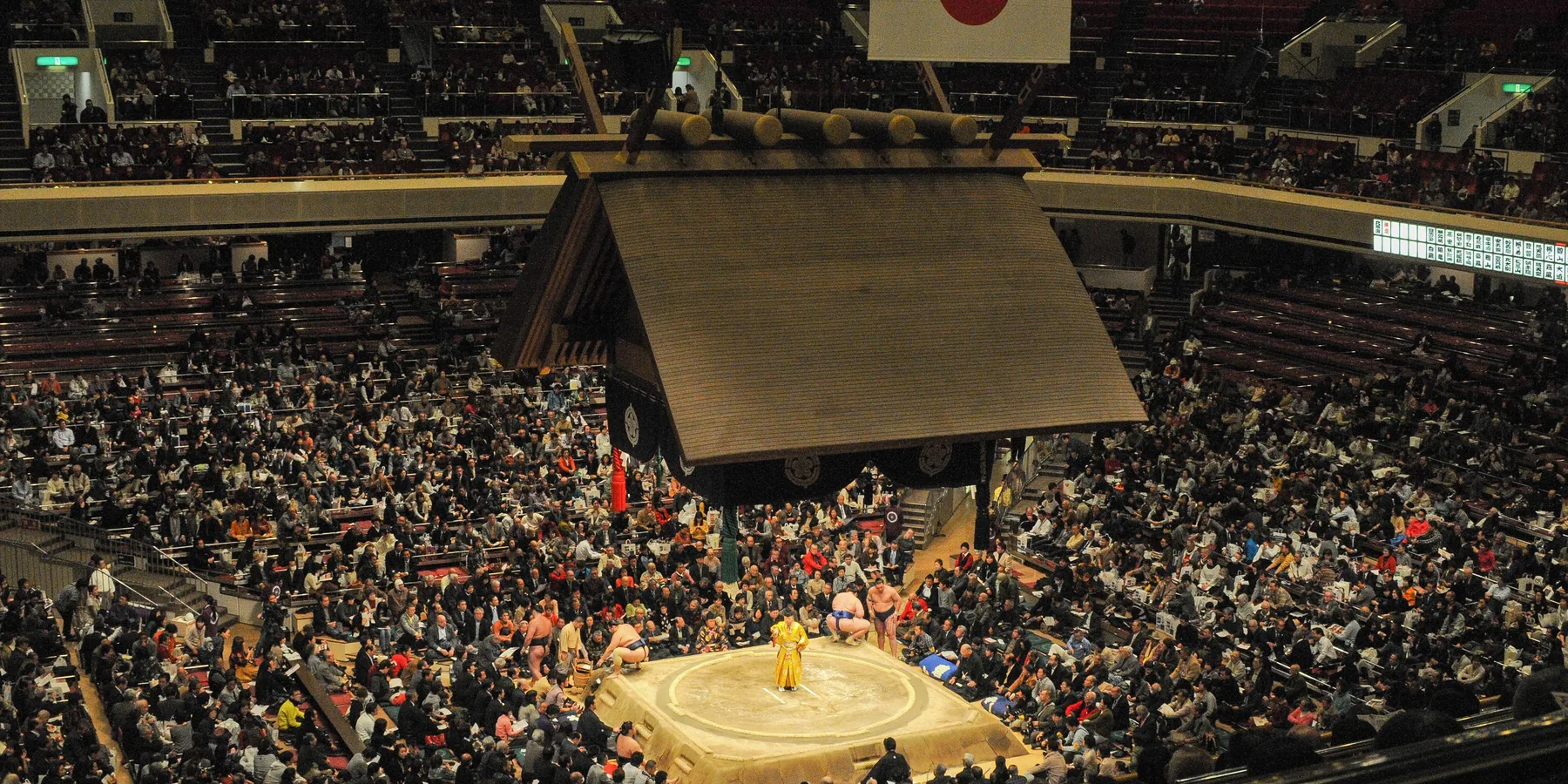 Přeplněná sumo aréna v Japonsku pohledem shora, s visící střechou ve stylu svatyně nad vyvýšeným ringem