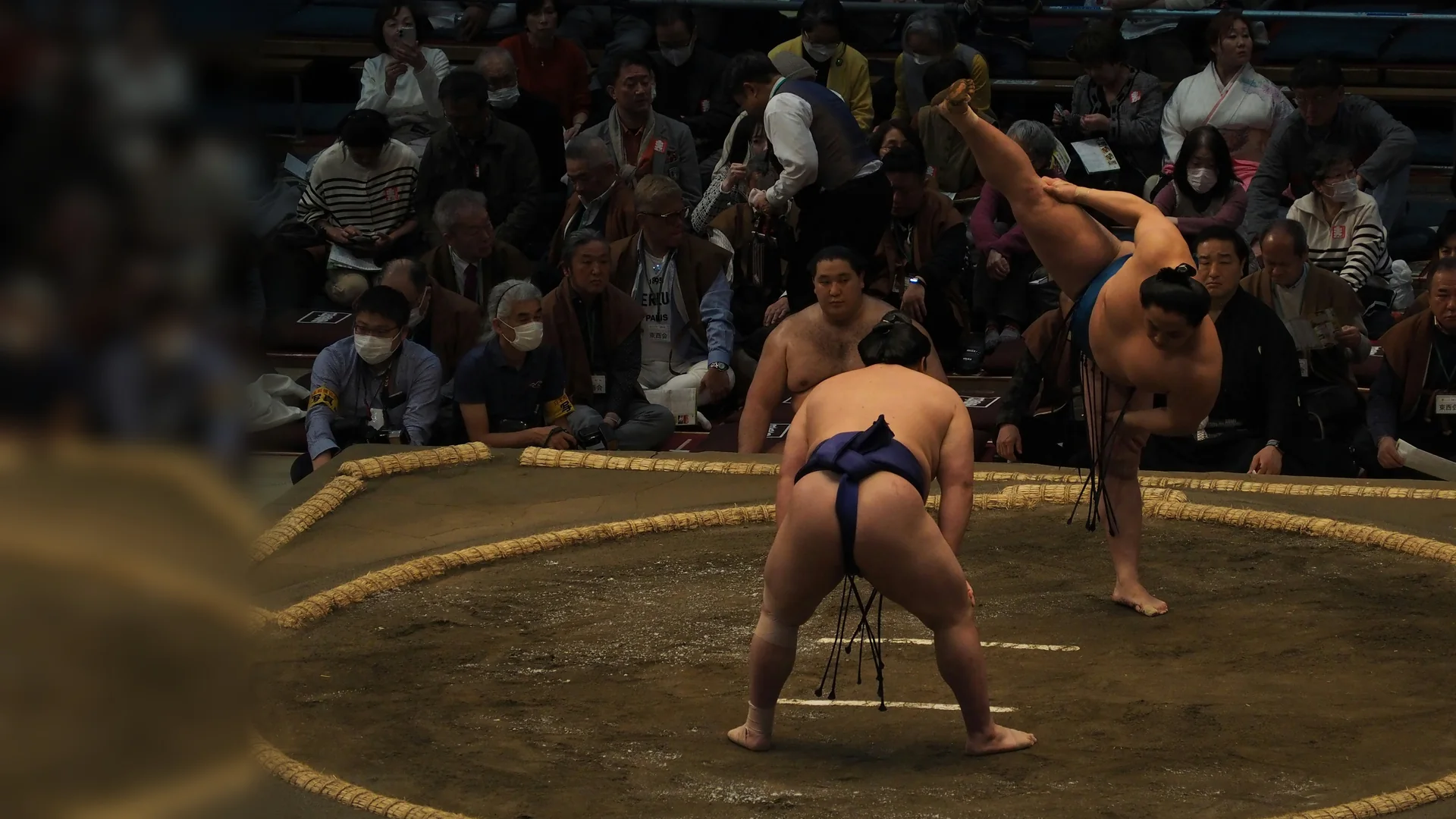 Dva sumo zápasníci stojí proti sobě uvnitř ringu z rýžových balíků, jeden s nohou zvednutou v rituálním dupnutí, zatímco diváci sedí těsně kolem hliněné plochy